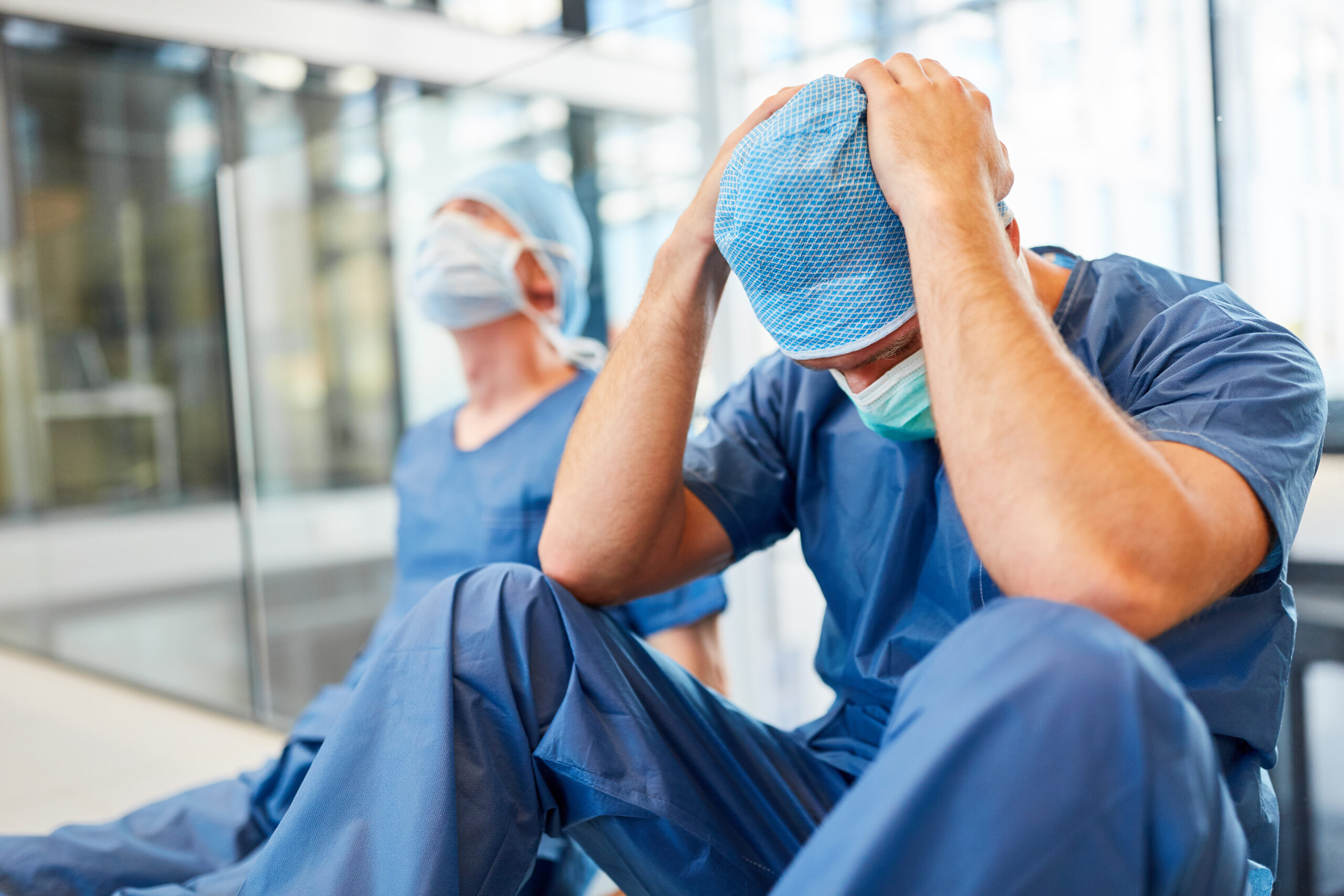 An exhausted surgeon in scrubs sits with his head in his hands while another surgeon stands nearby.