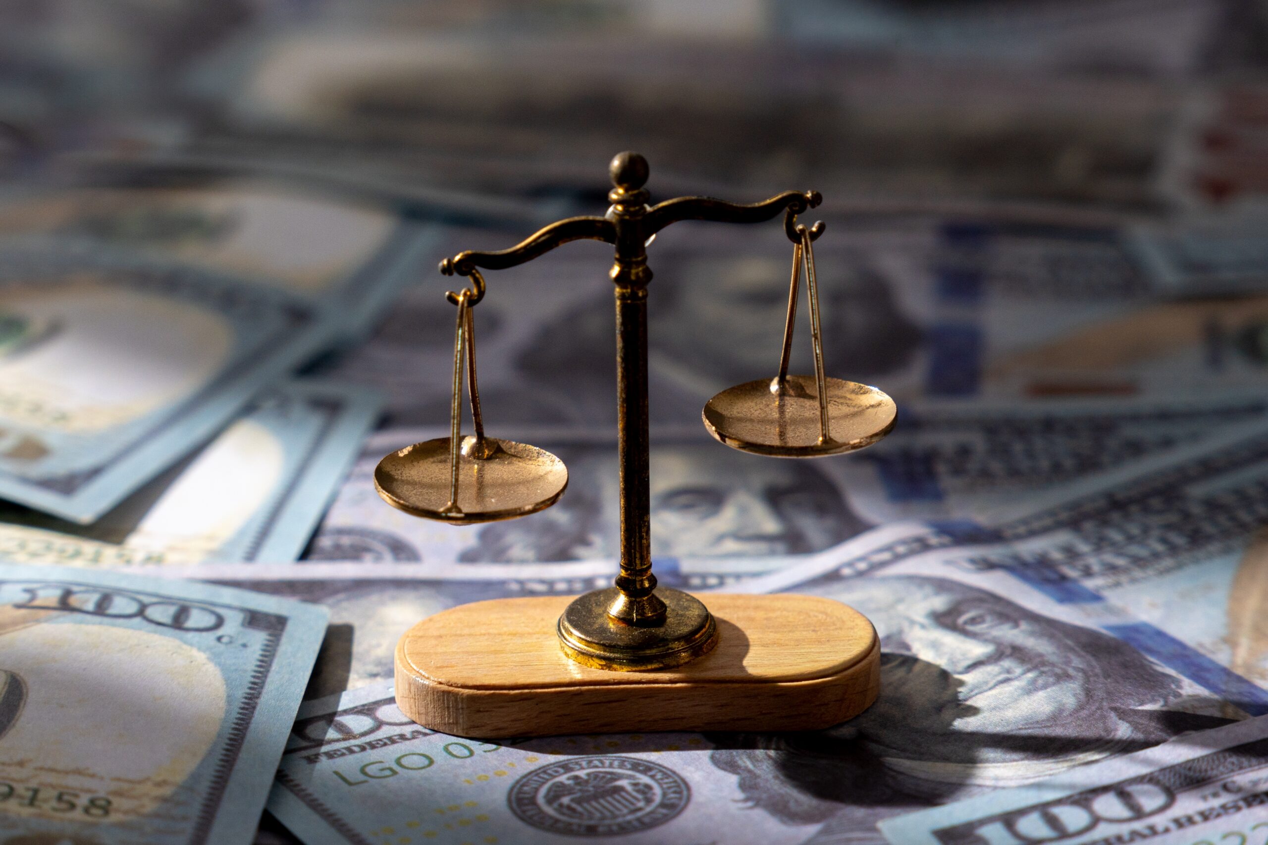 A small scale of justice balances on a wooden base atop scattered hundred-dollar bills.